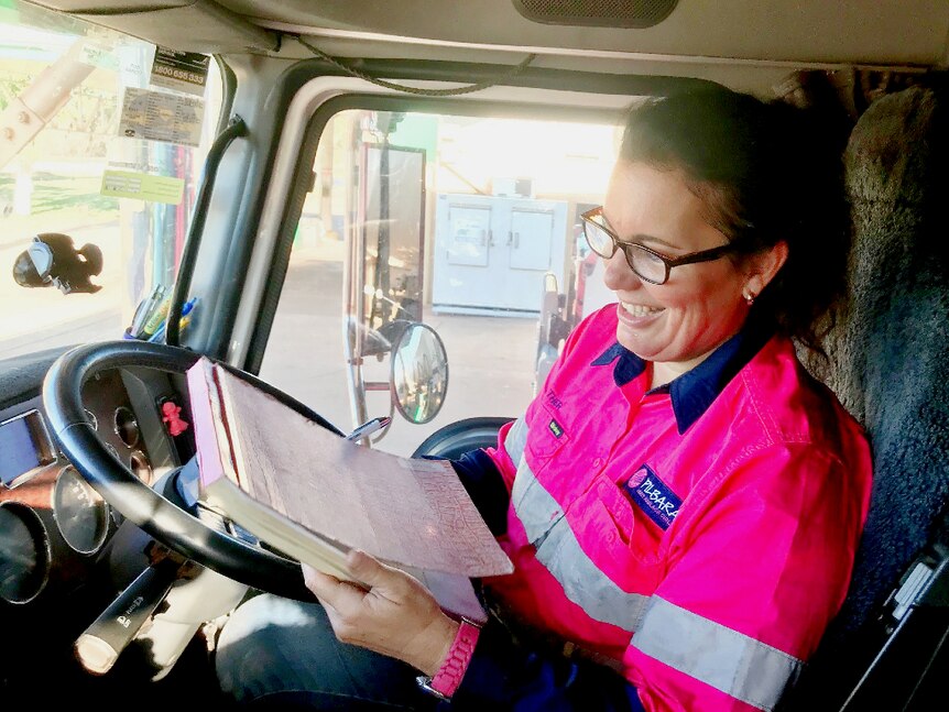 A female truck driver sits in the cabin and fills out the compulsory log book, she's smiling.