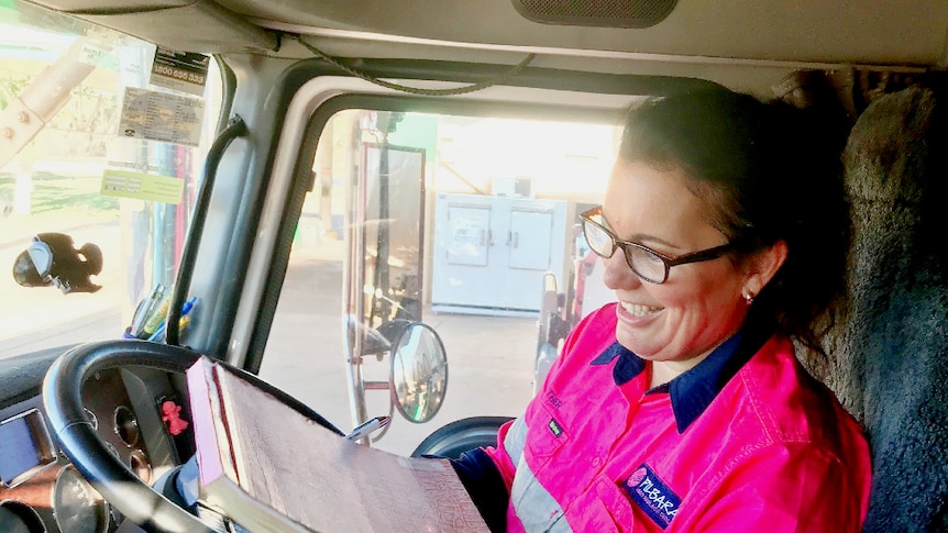 A female truck driver sits in the cabin and fills out the compulsory log book, she's smiling.