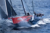A yacht with a red and blue hull crashes through a wave