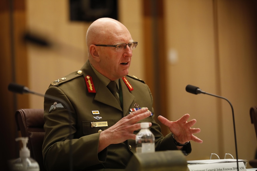 A man in military uniform sits at a committee bench, gesturing as he speaks.