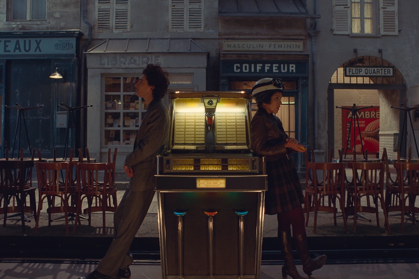 Two dressed-up 20-somethings - one girl, one boy - lean on either side of a cigarette machine in an outdoor bar in France