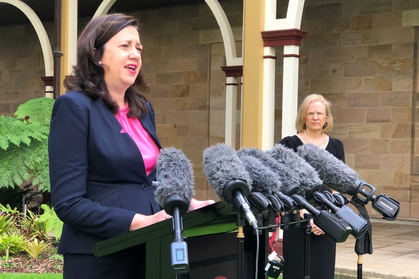 Annastacia Palaszczuk speaks at a podium surrounded by microphones and the Chief Health Officer Jeannette Young watches on