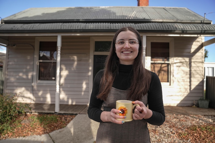 A young woman wearing grey dress over black skivvy stands in front of a house, smiling with a mug in her hands