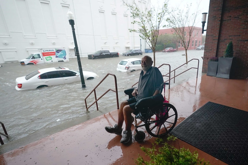 A man sitting in a wheelchair as waters rise, cars half submerged.