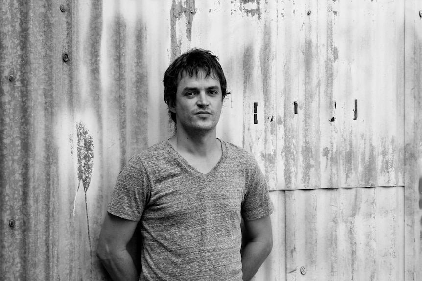 A black and white photo of a man standing next to a corrugated iron fence.