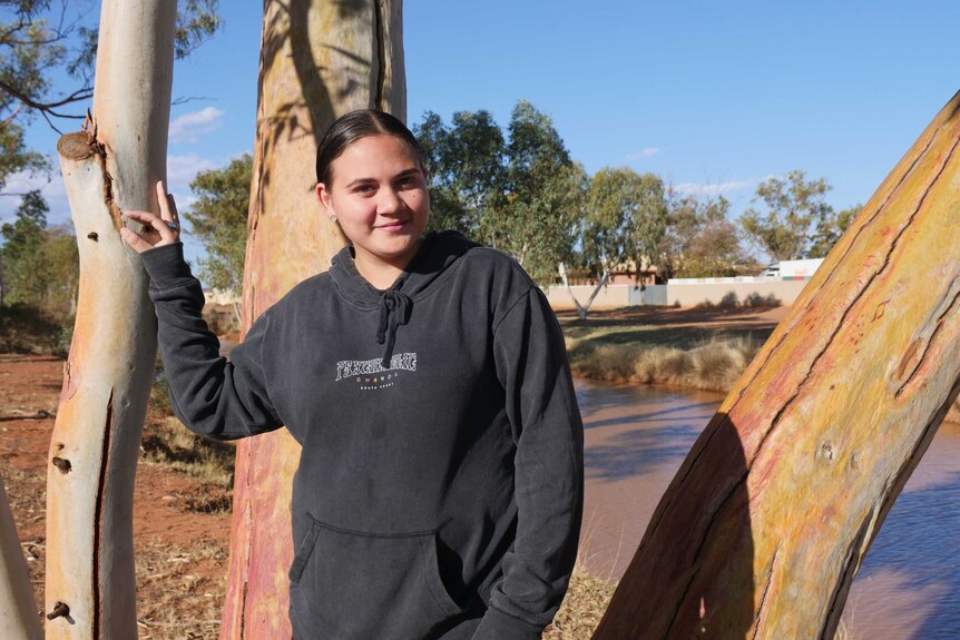 A young woman, smiling, stands next to a tree trunk