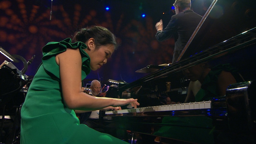 Andrea Lam plays grand piano in front of an orchestra. She's hunched over the piano with a look of intense concentration.