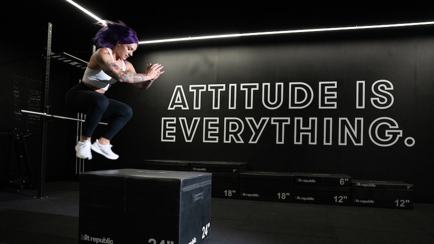 A woman with purple hair doing a box jump in front of a wall that reads "attitude is everything".
