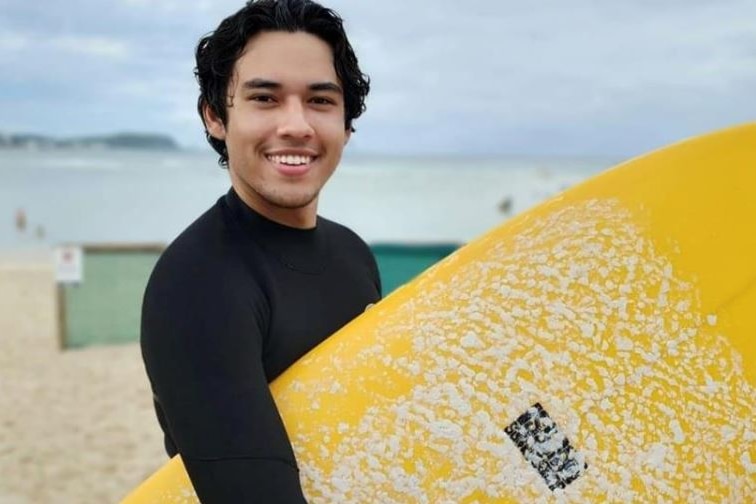 A smiling young man holding a surfboard on a beach.