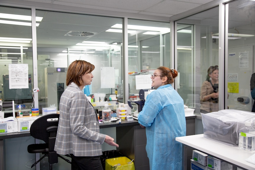 A woman in a chequered jacket speaks with a woman in scrubs at a science laboratory.
