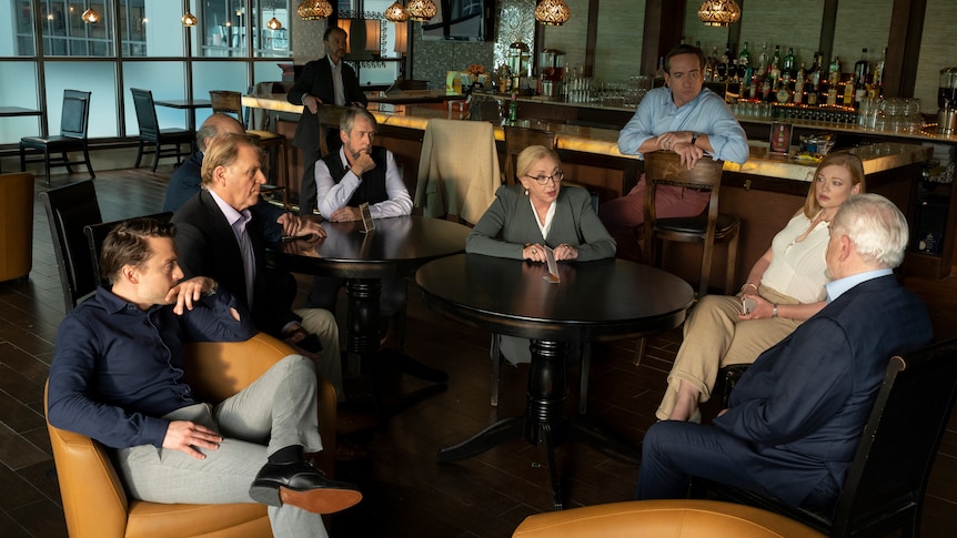 Characters of HBO show Succession sit in discussion in an empty bar, wearing business attire
