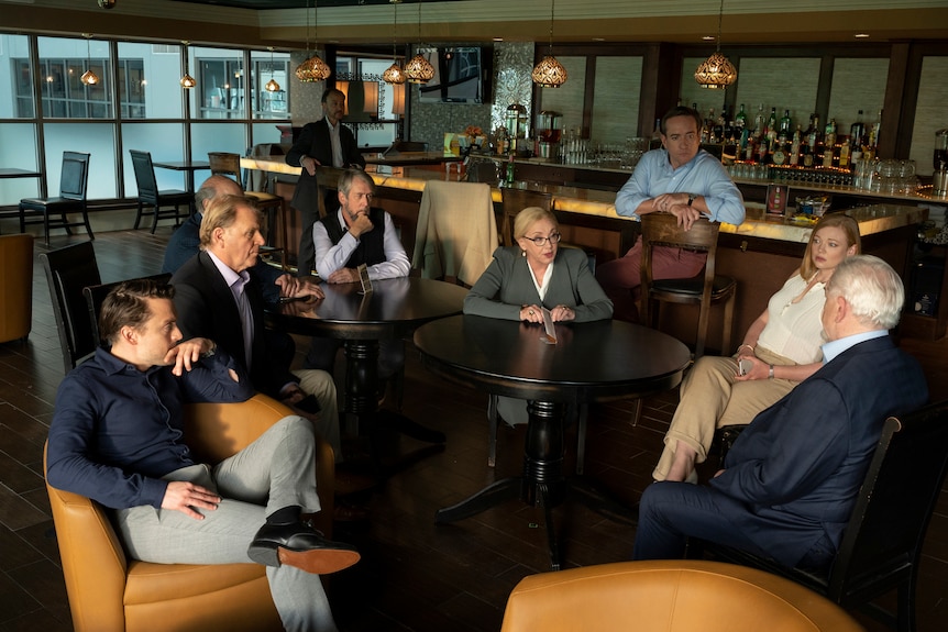 Characters of HBO show Succession sit in discussion in an empty bar, wearing business attire