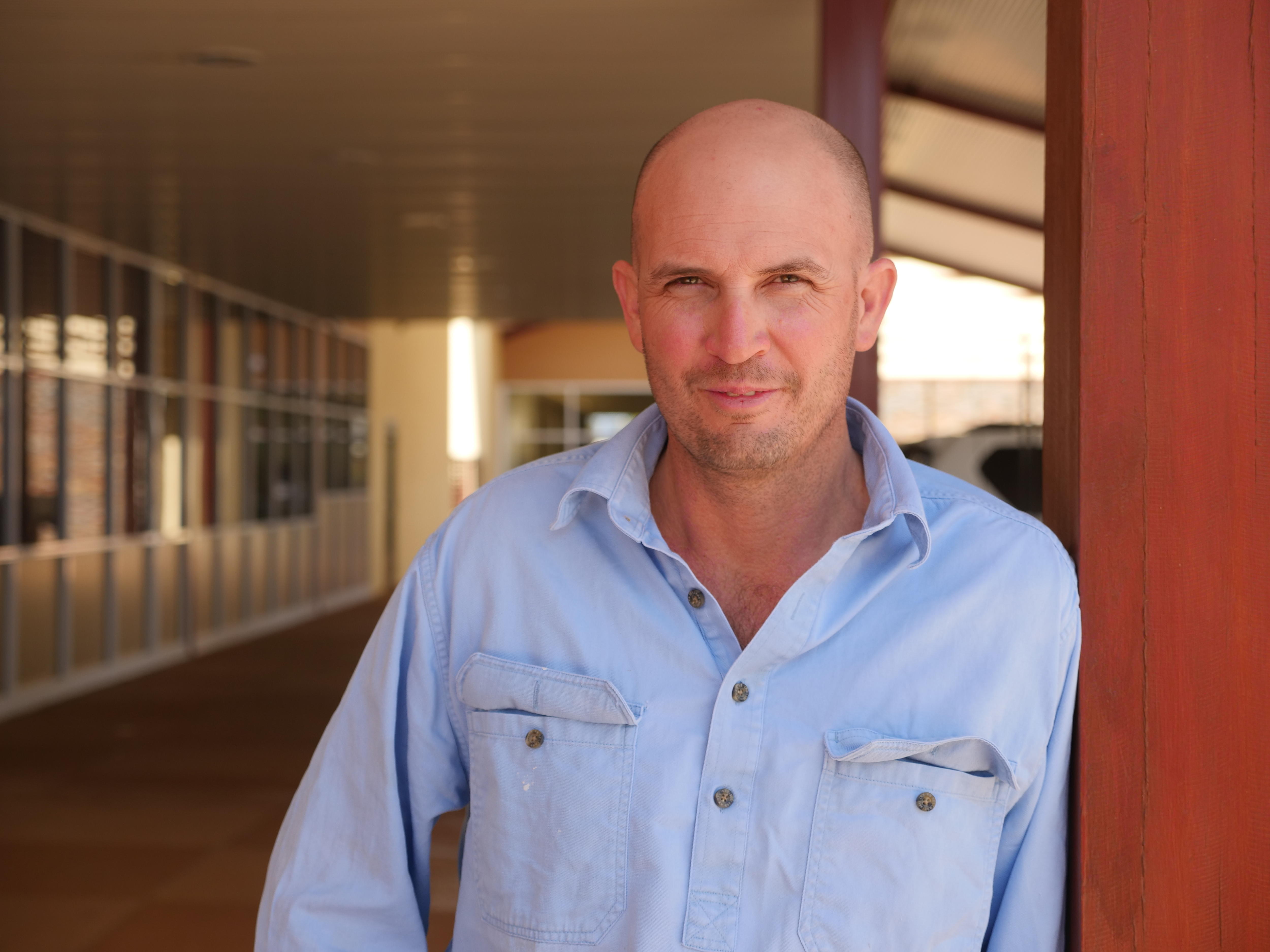 A bald man in a blue shirt stands in the shade, leaning on a pole.