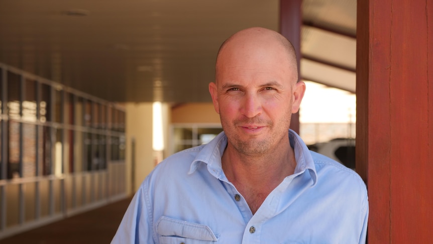 A bald man stands in the shade, leaning on a pole.