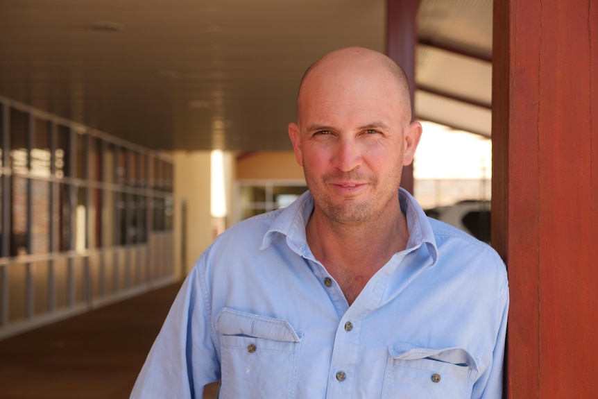 A bald man stands in the shade, leaning on a pole.