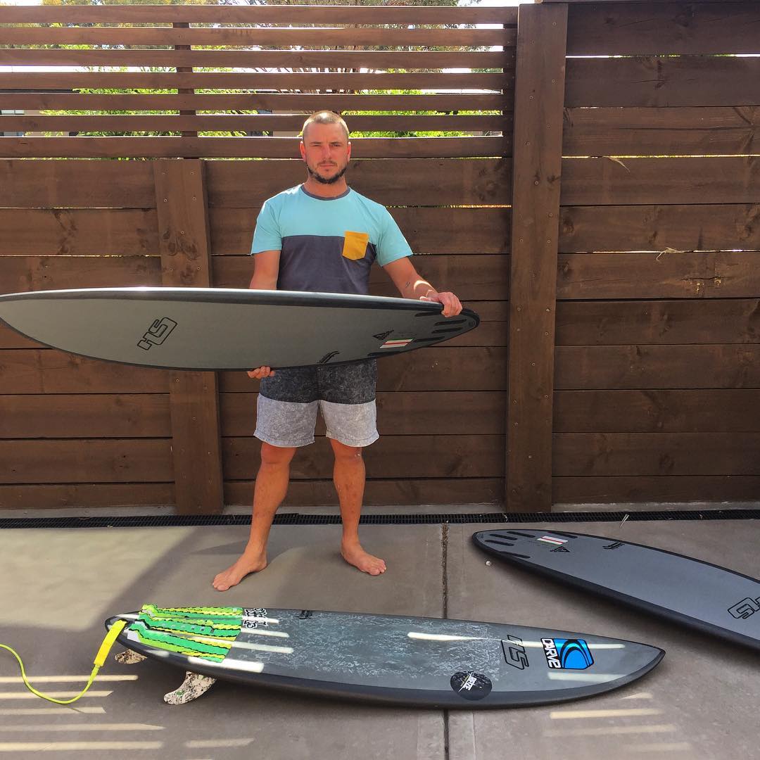 Man holding a surfboard, with two other surfboards near his feet