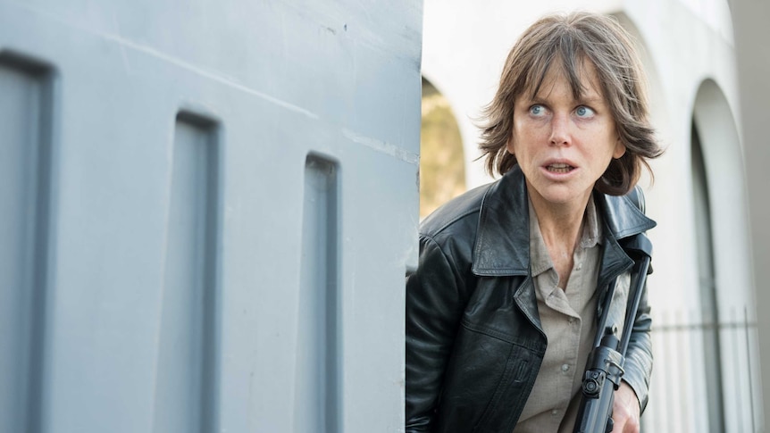 The actor crouches behind a wall, holding an assault rifle, looking out of frame warily.