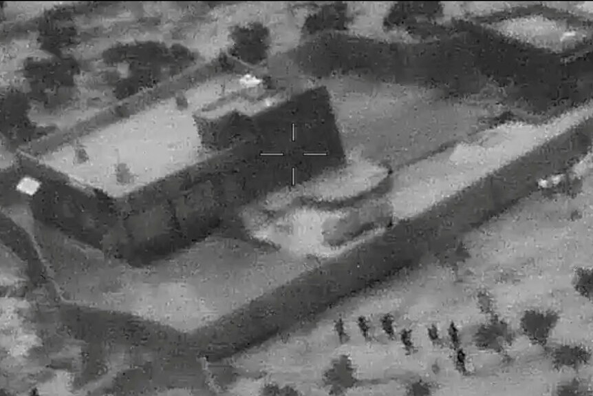 A black-and-white night-vision photograph, taken from the air, shows a small group of dark figures outside a walled compound.