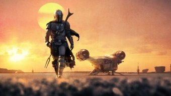 A still from the series Mandalorian, with the main character standing in front of a setting sun.