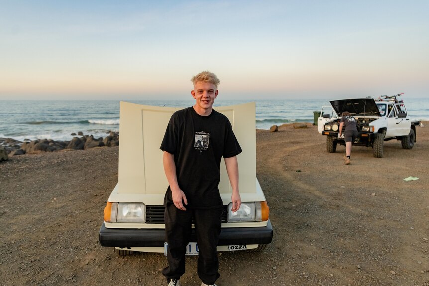 Teenage boy smiles while standing in front of a car by the ocean with clear blue skies.