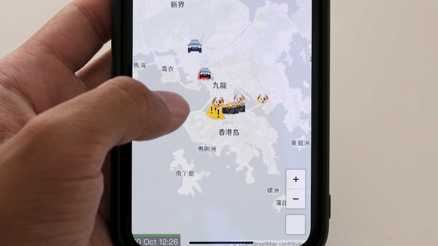 A hand holds a phone displaying the HKmap.live app. It shows a map of Hong Kong with emojis over points of interest.