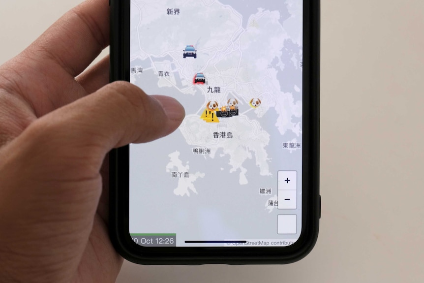 A hand holds a phone displaying the HKmap.live app. It shows a map of Hong Kong with emojis over points of interest.
