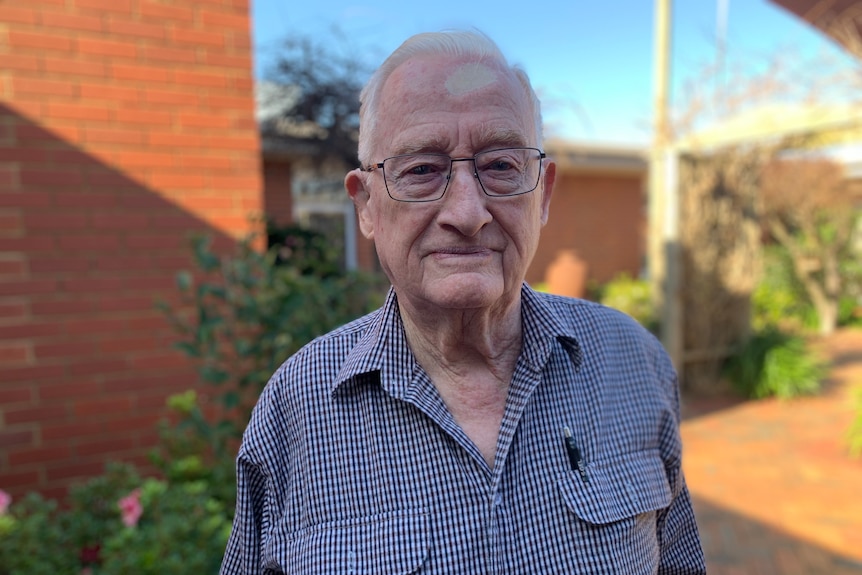 Elderly man wearing a blue and white check shirt and glasses, standing outside a building.