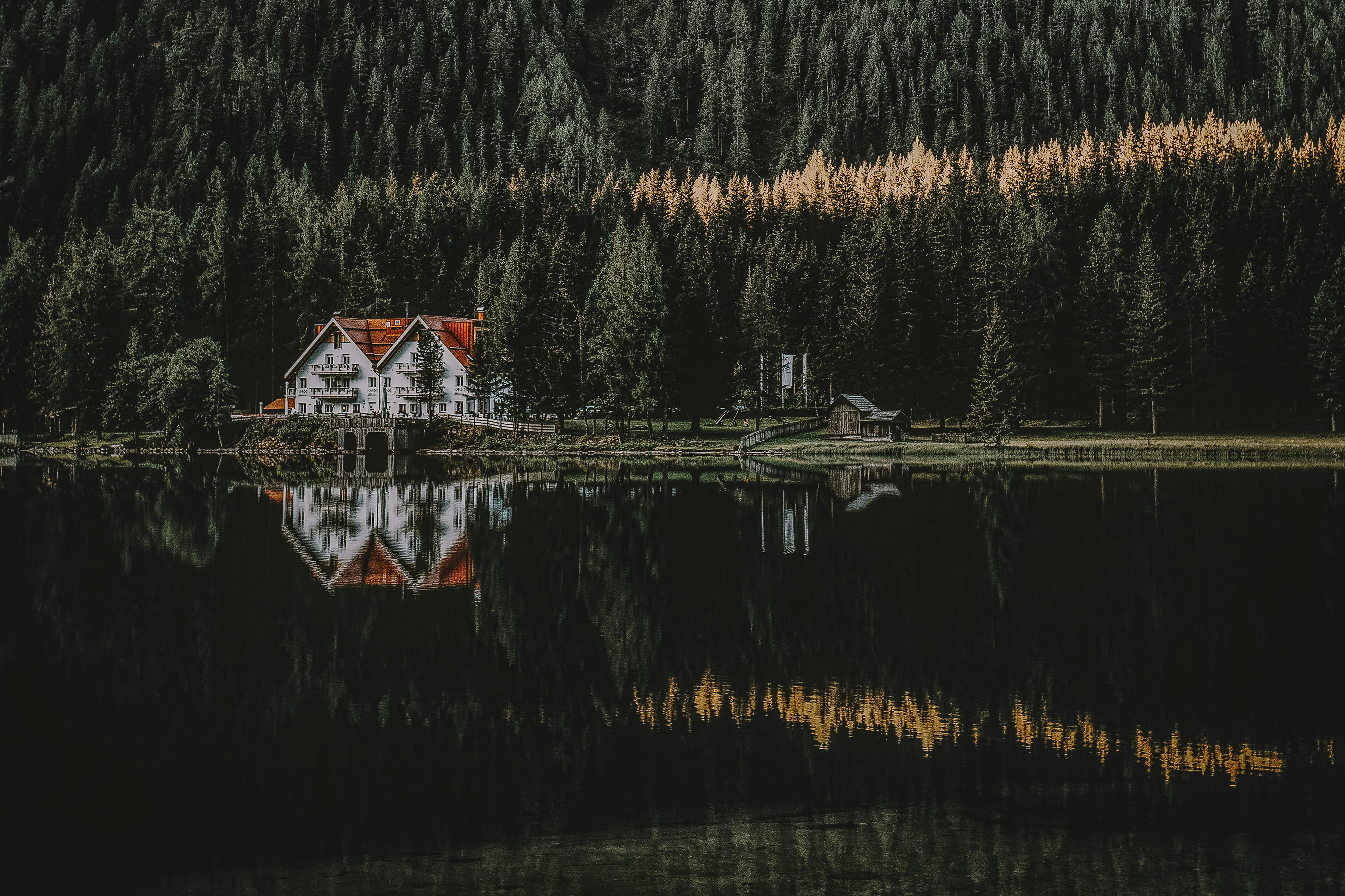 A white house with a red pitched roof sits on the edge of a lake surrounded by tall pine trees