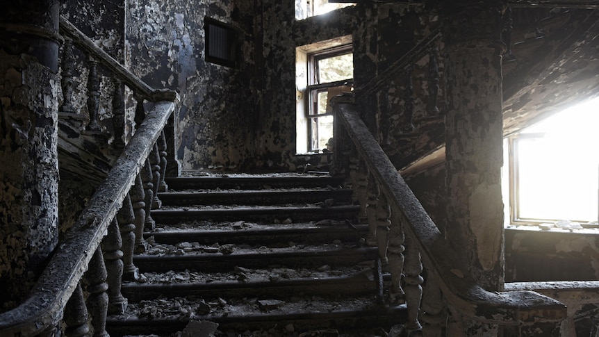 An ornate interior staircase and the walls around it are burned black, the stairs littered with debris