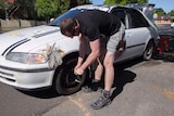 Good generic: A man bent over changing a tyre on a car that has dents patched with electrical tape