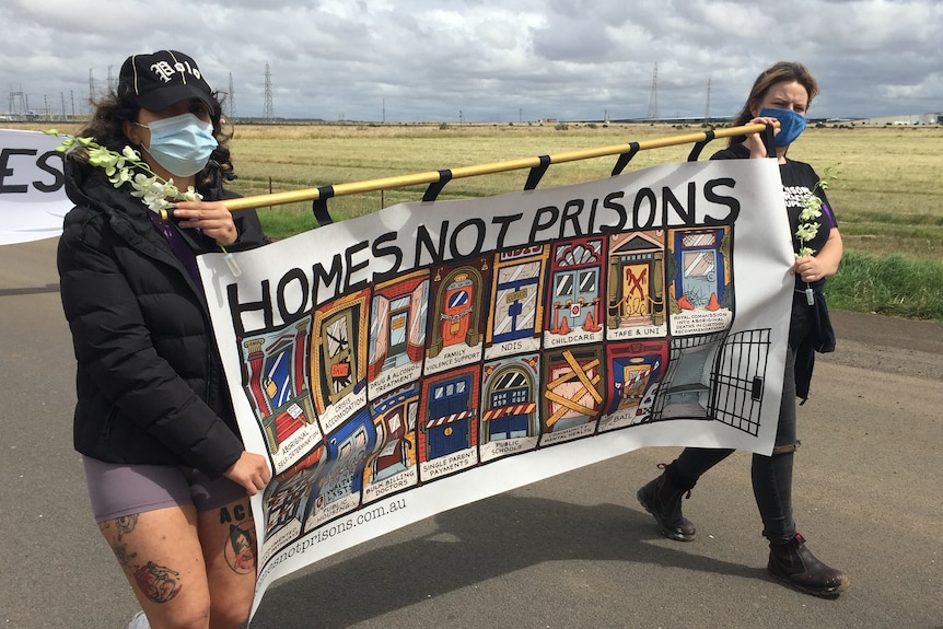 Two protesters march along a road holding an illustrated poster that reads 'HOMES NOT PRISONS'.