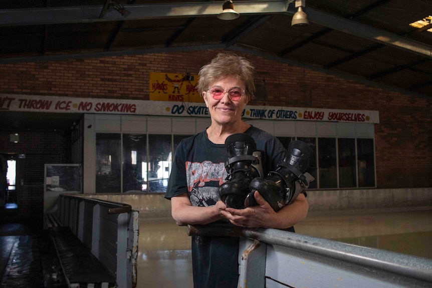 A woman holds a pair of ice skates while standing in front of an ice rink.