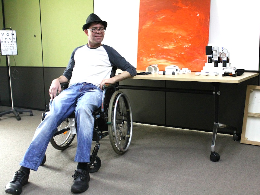 Man sits in a wheelchair with hat, gently smiling at camera in front of large orange-painted canvas, eye chart nearby.