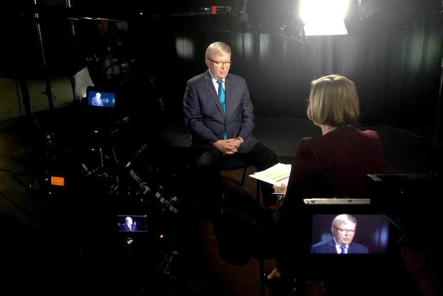 Man in television studio surrounded by lights and camera with woman sitting opposite.