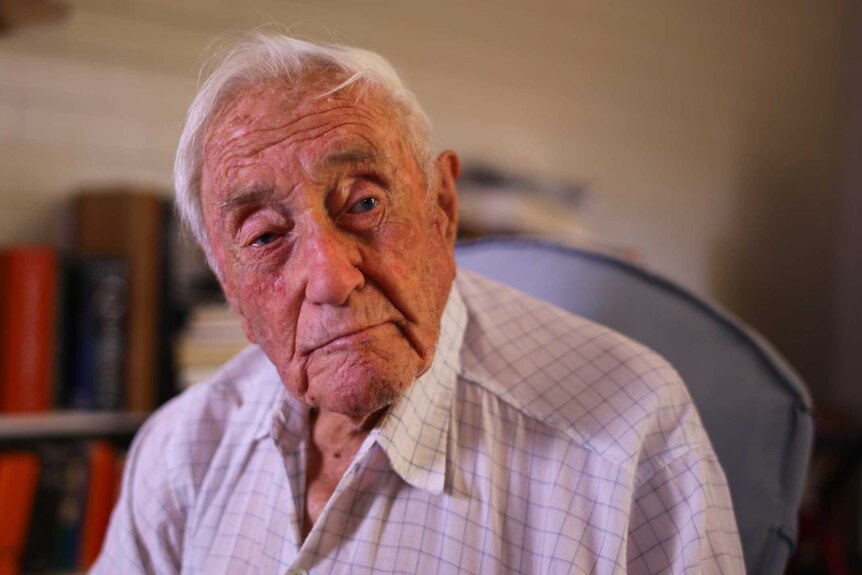 An elderly man sits on a chair in his home with a distant expression.