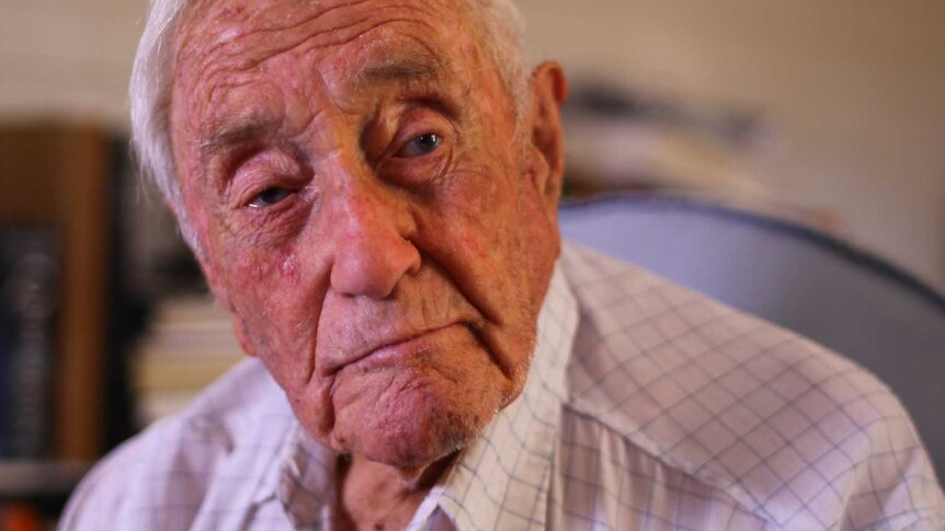 An elderly man sits on a chair in his home with a distant expression.