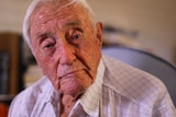 An elderly man sits on a chair in his home with a distant expression.