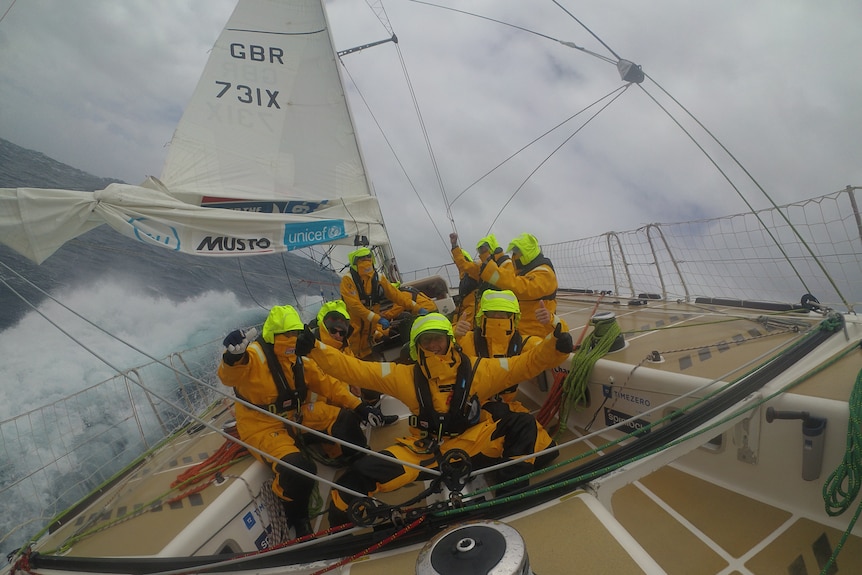 Six sailors cheer as their yacht races through rough seas. 