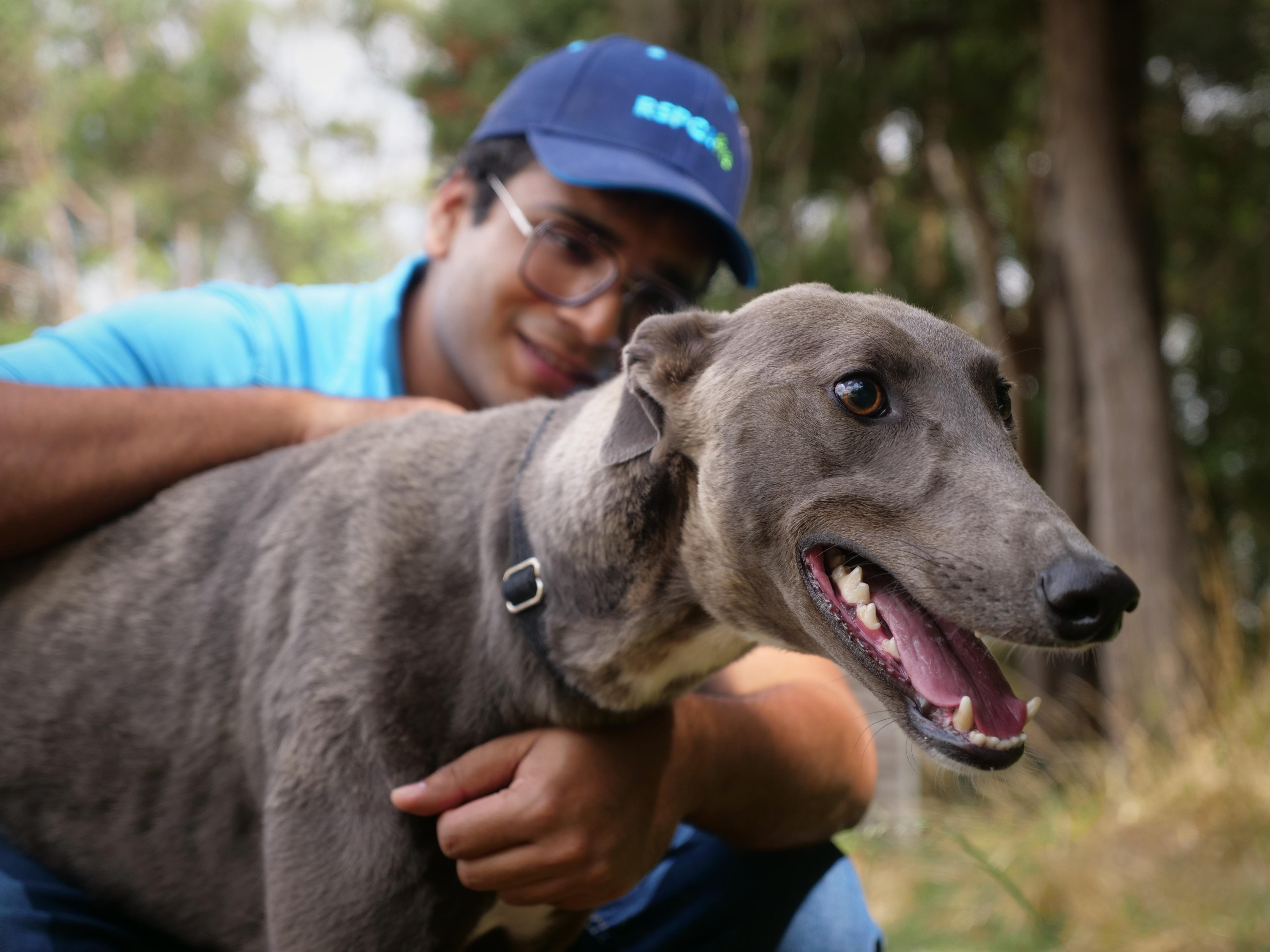 A man wearing a cap and glasses smiling as he hugs a grey greyhound, who is in the front of the shot.