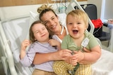 Grace, with blonde bun, smiles while lying in hospital bed cuddling her two young smiling