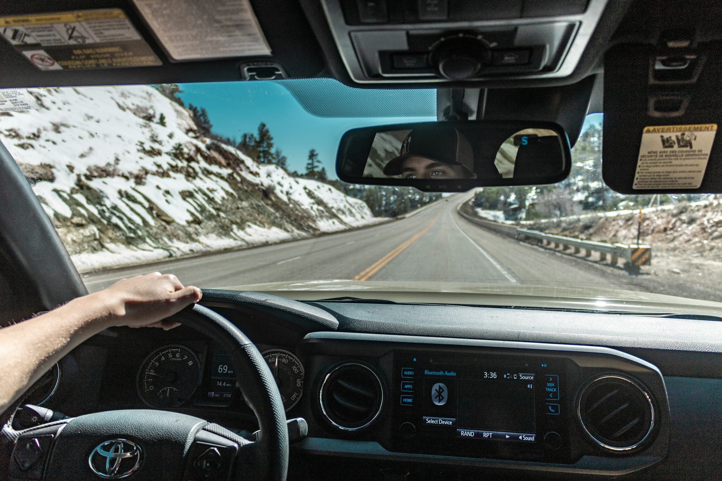 A photo taken inside a Toyota, showing a man's arm on the steering wheel as it drives down a road between snow-capped hills