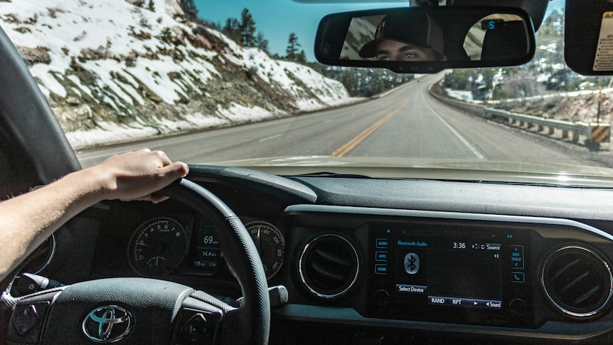 A photo taken inside a Toyota, showing a man's arm on the steering wheel as it drives down a road between snow-capped hills