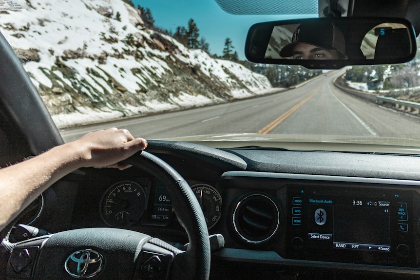 A photo taken inside a Toyota, showing a man's arm on the steering wheel as it drives down a road between snow-capped hills