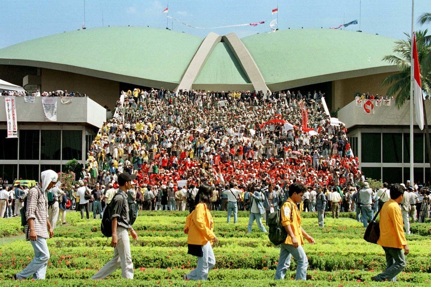 You view five students in bright shirts walking single file in front of a massive crowd protesting on tiered steps.