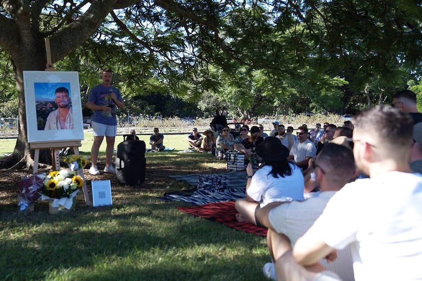 A group of people sit under a tree facing a man who is speaking and a large framed photo of Luke Davies.