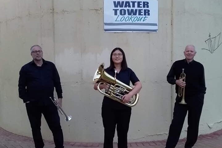 Three brass players stand together holding instruments in front of Berri water tower.