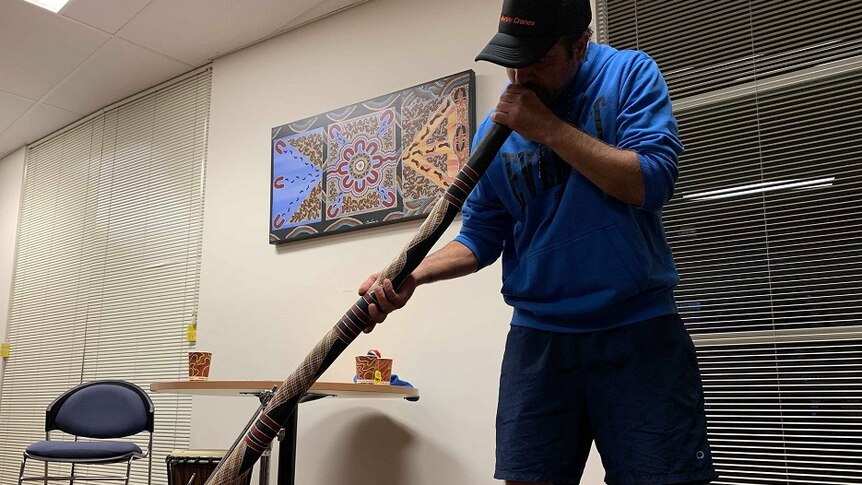 A man is standing in a bright room blowing into a didgeridoo. He is wearing a black cap with a blue jumper and blue shorts.