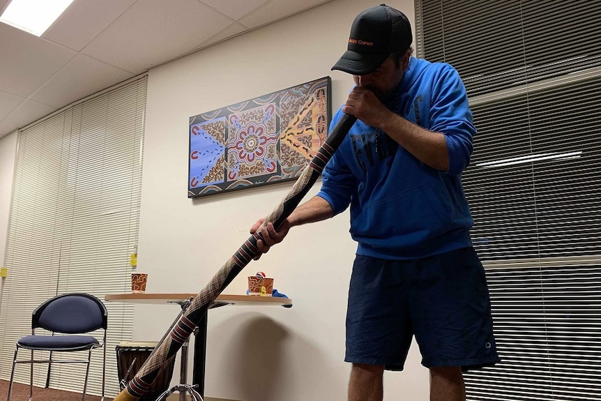 A man is standing in a bright room blowing into a didgeridoo. He is wearing a black cap with a blue jumper and blue shorts.