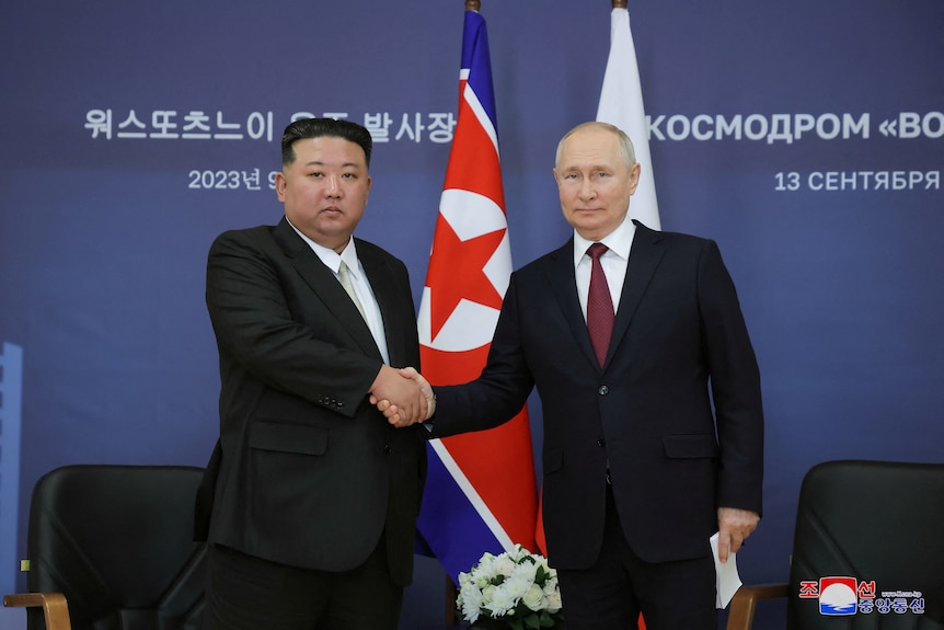 Kim Jong Un and Vladimir Putin shake hands in front of two flags.