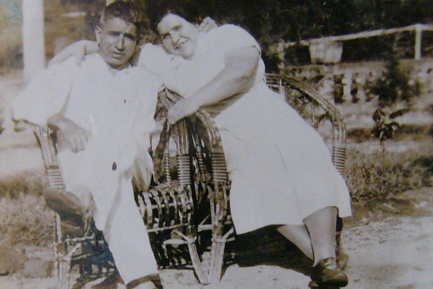An archive photo of a middle-age man and woman embracing oneanother while sitting on wicker chairs.